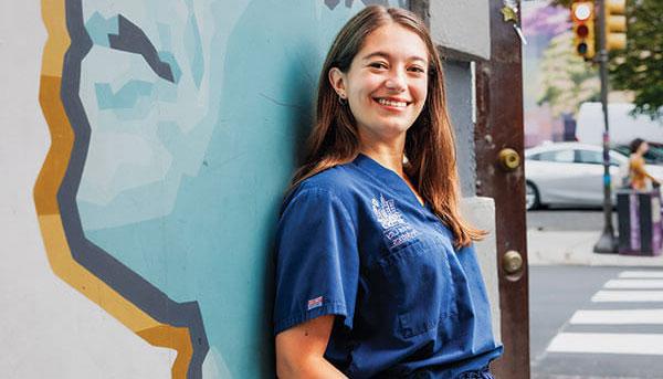 Jessica Rose ’16 wearing blue scrubs leaning against wall